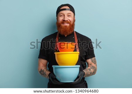 Satisfied male cook holds pile of colorful kitchen utensils, uses dish for cooking dinner, invites you in world of tastes, wears black uniform, rubber protective gloves, isolated on blue background
