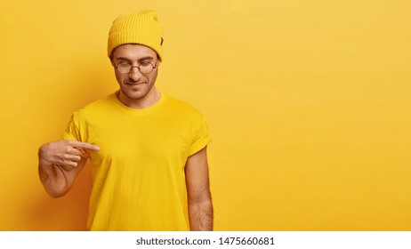 Satisfied Male Model Points At Blank Space Of His Yellow T Shirt, Shows Mockup Place For Slogan, Advertisement Or Brand Label, Focused Down, Advertises Clothes, Likes Yellow Color, Isolated.