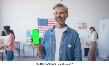 Satisfied man holding smartphone with green screen in one hand. Chroma key visible on phone display. Voters electing for Presidential Candidate in background. American flag hanging on wall. - Powered by Shutterstock