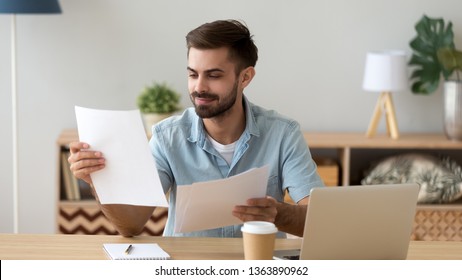 Satisfied man reading paper documents, letters, receiving pleasant news, working with laptop at workplace, sitting on desk, successful businessman, freelancer looking at contract with good offer - Powered by Shutterstock