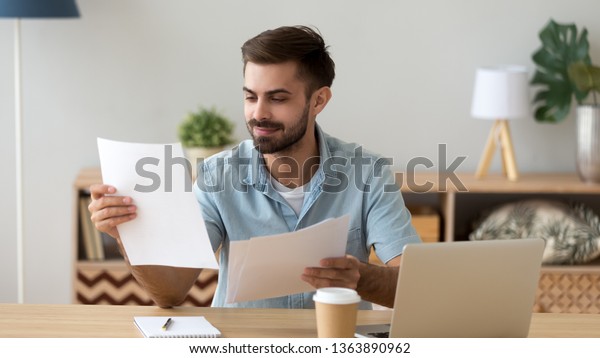 Satisfied man reading paper documents, letters,\
receiving pleasant news, working with laptop at workplace, sitting\
on desk, successful businessman, freelancer looking at contract\
with good offer