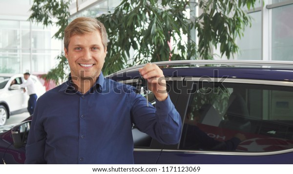 A satisfied man is standing by the new car. He\
bought it. He shows the keys of this automobile. He looks very\
pleased and happy