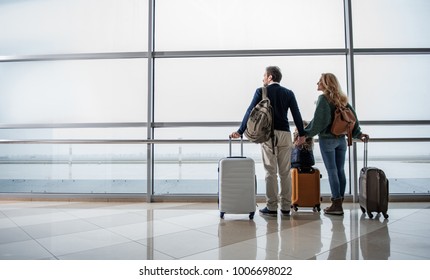 Satisfied man, woman and girl looking out from big window at the airport. They are taking pleasure in airplanes moving along the runway. Copy space in left side