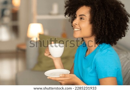 Satisfied millennial african american curly lady enjoy relax, cup of tea on sofa, look at copy space in cozy living room interior. Good morning, coffee break and silence, evening relax alone