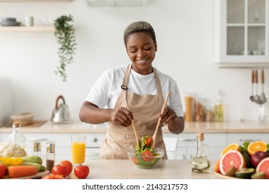 Satisfied millennial african american lady in apron preparing organic vegetable salad at table in modern kitchen interior. Food blog, health care and cooking lunch at home during covid-19 quarantine - Powered by Shutterstock