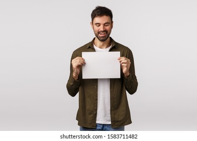 Satisfied, pleased charming handsome caucasian man with beard in modern outfit, have something to offer, holding blank piece paper, recommend visit web page, look at writing with pleased smile - Powered by Shutterstock