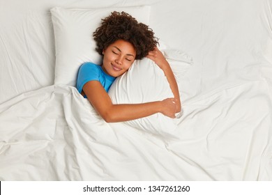 Satisfied Pretty Afro American Young Woman Has Peaceful Sleep, Lies In Bed On Comfortable Pillow, Sees Pleasant Dreams, Has Gentle Smile, Enjoys Softness Of Bedclothes, Being Under White Blanket