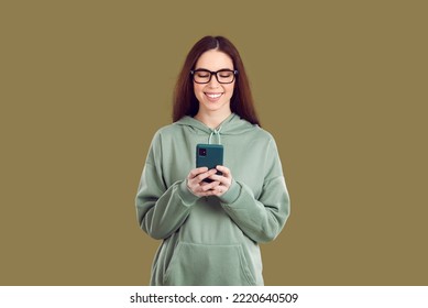 Satisfied Pretty Young Caucasian Woman Holding Mobile Phone In Front Of Using Dating Apps Or Social Networks To Communicate With Boyfriend Wearing Casual Sweatshirt Stands In Green Studio