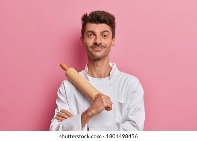 Satisfied Professional Cook Holds Wooden Rolling Pin, Going To Prepare Dough For Pizza, Dressed In White Uniform, Stands Against Pink Background. Cooking, Restaurant Staff, Profession Concept