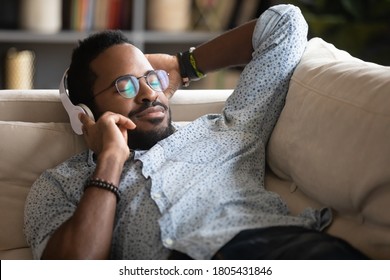 Satisfied relaxed African American man wearing headphones enjoying favorite music close up, pleasant sounds with closed eyes, young male wearing glasses lying on cozy couch at home, daydreaming