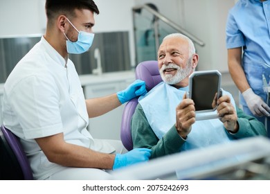 Satisfied Senior Man Looking His Teeth In A Mirror And Talking To His Orthodontist After Dental Appointment At Dentist's Office. 