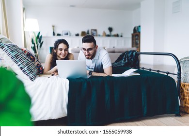 Satisfied Smiling Amorous Couple Having Rest On Comfy Vintage Bed While Using Laptop In Modern Light Bedroom On Blurred Background