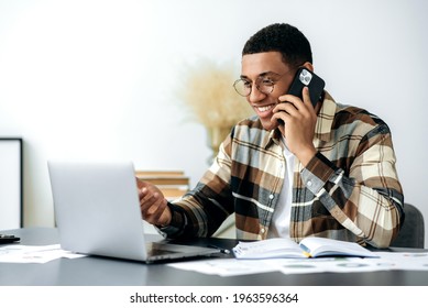 Satisfied Smiling Mixed Race Latino Male Freelancer Or Manager With Glasses, Have Cell Phone Conversation, Sits At Work Desk, Uses Laptop, Checking E-mail, Looking For Information At Internet