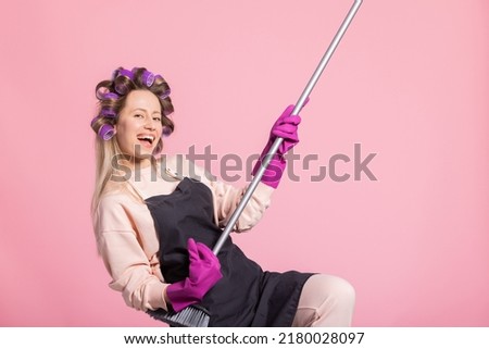 Satisfied smiling woman with long blonde hair curled rollers on head has protective rubber cleaning gloves on hands holds a broom pretending to play guitar fooling around.