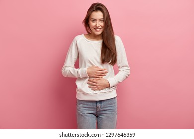 Satisfied smiling young woman keeps both hands on belly, being in good mood after eating delicious supper, demonstrates she is full, isolated over pink background. Pleasant feeling in stomach - Shutterstock ID 1297646359