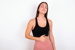 Satisfied Smiling Young Woman Wearing Sportswear Over White Studio Background , Keeps Hands On Belly, Being In Good Mood After Eating Delicious Supper, Demonstrates She Is Full. Pleasant Feeling In St