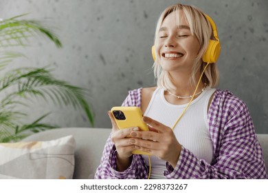 Satisfied smiling young woman wearing casual clothes headphones listen music use mobile cell phone sits on grey sofa couch stay home hotel flat rest relax spend free spare time in living room indoor
