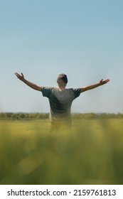 Satisfied successful farmer raising hands in victorious pose in unripe barley crops field on sunny spring day. Rear view of farm worker wearing green t-shirt and trucker hat with arms lifted up over - Shutterstock ID 2159761813