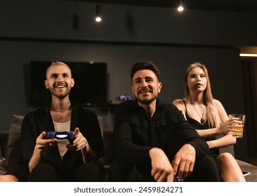 Satisfied with virtual match young men hold joysticks sitting on cozy sofa. Blonde woman savors cold beer resting with friends - Powered by Shutterstock