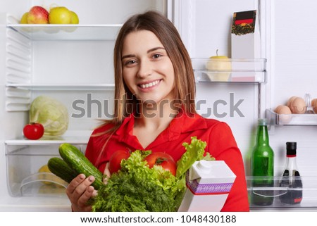 Satisfied woman holds fresh vegetables and milk, going to put them in refridgerator, has broad smile and happy expression, being vegeterian, eats only healthy food. People and nutrition concept