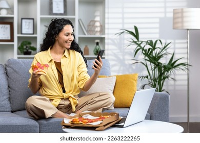 Satisfied woman at home relaxing sitting on sofa in living room and eating pizza, Hispanic woman ordered food online using app on phone, satisfied with fast delivery. - Powered by Shutterstock