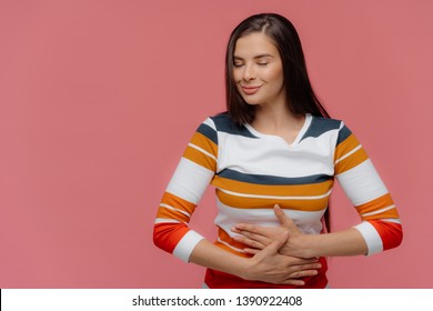 Satisfied Woman Keeps Hands On Belly, Feels Replete After Delicious Supper, Has Full Stomach, Long Hair, Appealing Appearance, Wears Jumper, Models On Pink Wall. Lady Finds Out About Pregnancy