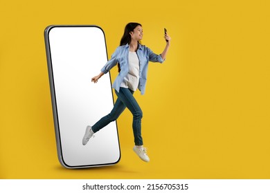Satisfied young asian lady jumping off huge phone with blank screen froze in air and look at smartphone isolated on yellow background, full length. Selfie for blog and social networks, modern device