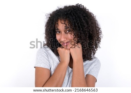 Satisfied Young beautiful girl with afro hairstyle wearing grey t-shirt over white background touches chin with both hands, smiles pleasantly, rejoices good day with lover