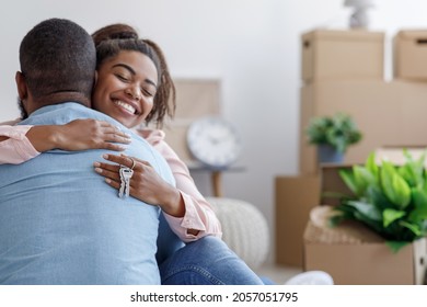 Satisfied young black family hugging, hold key and enjoy buying real estate in room with cardboard boxes and plants. Couple excited by relocation in new house. Move day, new life, housing improvement