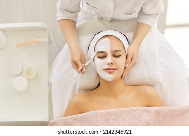 Satisfied Young Caucasian Beautiful Woman Uses Services Of Professional Beautician In Beauty Salon Or Spa. Unrecognizable Beautician Applied White Mask To Half Of Client's Face With Brush. Top View.