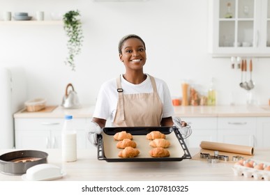 Satisfied Young Cute Black Woman In Apron Shows Ready Croissants In Modern Kitchen Interior. Housewife Make Homemade Eat, Food Blogger Cook Pastries At Home. Prepare Tasty Diner For Family, Free Space