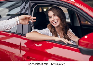 Satisfied young female sitting in new red automobile and receiving keys from unrecognizable salesman in car salon