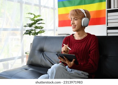 Satisfied young gay queer man in headphone and red sweater using digital tablet in living room with pride rainbow flag on background