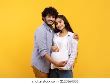Satisfied young indian man hugging pregnant wife and touching belly, smiling together at camera over yellow background. Couple awaiting baby, cute parents-to-be enjoy tender moment - Shutterstock ID 2123864888