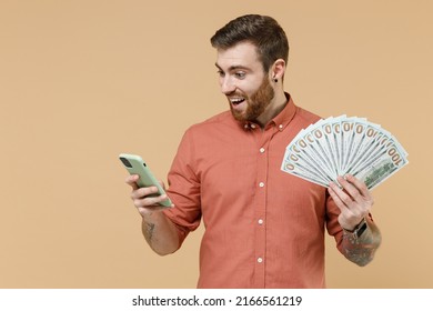 Satisfied young man 20s he wears orange shirt hold in hand mobile cell phone fan of cash money in dollar banknotes isolated on plain pastel light beige background studio portrait. Tattoo translate fun