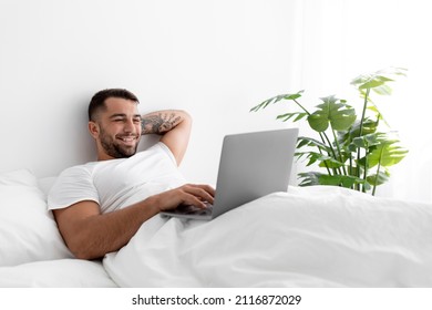 Satisfied young muscular caucasian man typing on computer lies on bed in bedroom interior at morning. Guy relax at home, watch funny video. Social media, video call, great offer, message and ad