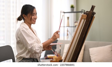 Satisfied Young Woman Painting With Watercolor On Canvas In Bright Living Room. Art, Creative Hobby And Leisure Activity Concept