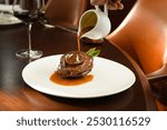 Sauce cascading from a white jug onto a juicy steak, garnished with herbs on an elegant plate, with a wine glass in the background.