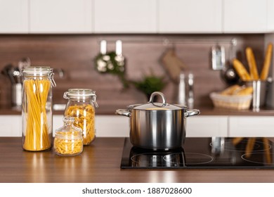 Saucepan On Electric Stove Close To Dry Uncooked Pasta In Glass Jar. Contemporary Interior With White Kitchen Cupboards, Wooden Countertop And Induction Cooker