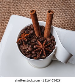 A Saucer With A White Cup On The Table. The Cup Contains Coffee Beans, Two Cinnamon Sticks And Two Star Anise.View From Above.Copy Space.