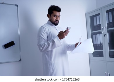 Saudi Arab Man Using Phone While Looking At Paper In Office         