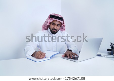 Saudi Arab man writing on page in diary while working on laptop    