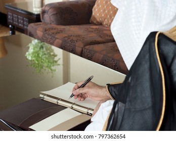 Saudi Arabian Man Hand Writing On A Notebook In A Luxury Home Environment, Wearing Saudi Thob, Ghutra And Black Bisht