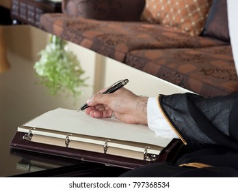 Saudi Arabian Man Hand Writing On A Notebook In A Luxury Home Environment, Wearing Saudi Thob, Ghutra And Black Bisht