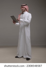 A saudi character wearing thob standing working on tablet on withe background.