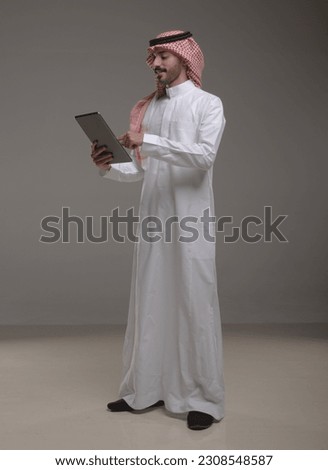 A saudi character wearing thob standing working on tablet on withe background.