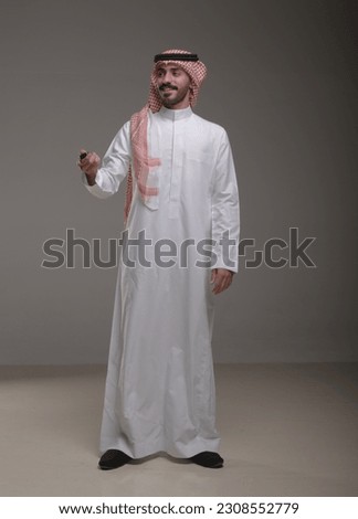 A saudi character wearing thob standing carrying a car key on withe background.