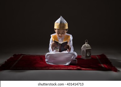 Saudi Kid in Traditional Saudi Uniform Sitting on Red Praying Mat and Reading Quran on Dark Background.