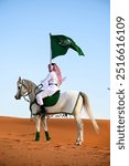 A Saudi man dressed in traditional white thobe and headscarf proudly holds the Saudi flag. He rides a majestic Arabian horse with grace. Celebrating National Day or founding day