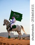 A Saudi man dressed in traditional white thobe and headscarf proudly holds the Saudi flag. He rides a majestic Arabian horse with grace. Celebrating National Day or founding day